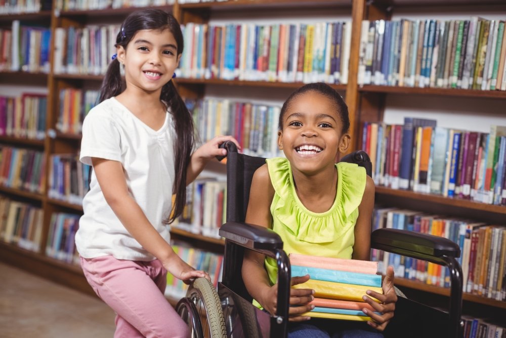 One child standing next to another in a wheelchair in a library